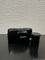 Front view of Olympus Mju Zoom camera with lens cover closed and film roll canister beside it