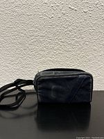 Black soft carrying case with zipper and shoulder strap placed on a flat surface against a beige textured wall.