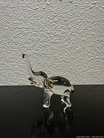 Front angle view of a small clear glass elephant figurine with gold accents on trunk, ears, and tusks placed on a black surface against textured white wall.