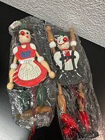 Front view of two vintage wooden pull string dolls wrapped in plastic, highlighting their painted faces, clothing, and shoes.