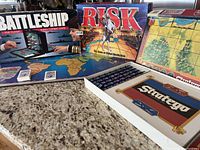 Photo showing three boxed classic board games - Risk, Battleship, and Stratego - along with game boards laid out on a surface.