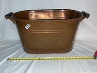 Front view of the large copper bin with black handles on a white background and a measuring tape showing approximately 25 inches in length.