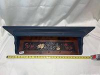 Top view of coat rack showing dark blue wooden frame, floral and butterfly inlay on reddish wood base, measuring tape showing approx 19.5 inches width.