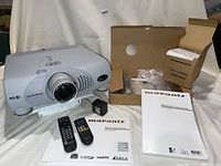 Marantz projector with lens, two remote controls, power adapter, user guides, and lamp unit box arranged on a white cloth.