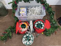 Wide view of entire lot showing the green garland surrounding two red storage containers with reels of white Christmas icicle lights and a large plastic bin with loose white string lights and two unopened boxes of mini clear icicle lights.