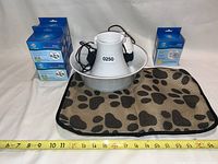 One photo showing the white pet bubbler fountain centrally positioned on the moisture-wicking mat with black paw prints, alongside blue boxes of replacement filters stacked on the left and one box on right.