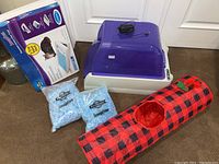 Photo shows the Petsafe automatic litter box cleaner with a purple translucent lid, two bags of PetSafe ScoopFree crystal litter, the 3-tray disposable litter tray box, and a red and black checkered crinkle cat tunnel.
