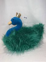 Front and side views of the Princess Peacock stuffed toy showing the details of the blue velvet head, yellow beak, black eyes, gold crown, and fluffy green faux fur tail.