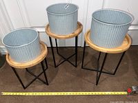 Three aluminum planters on wooden stands displayed together, measuring tape shows width of about 18 inches diameter for planter.