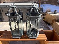 Pair of two antique metal and glass lanterns shown side by side on wooden bench outdoors in daylight.