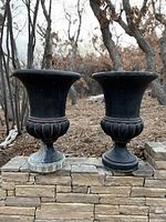 Pair of tall black garden planters sitting outdoors on stone with trees in background.
