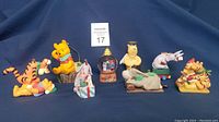 Shows the entire lot of eight Winnie the Pooh collectible ornaments arranged on blue background with a Maxsold auction card