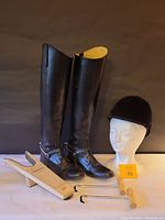 Pair of black tall leather riding boots, black velvet riding helmet on a mannequin head, wooden boot shaper and two metal hooks with wooden handles displayed on white surface