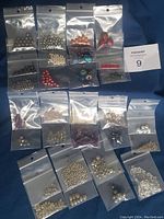 29 individual sealed bags of jewelry beads displayed on a blue cloth, showing various metallic, glass, and pearl-like beads in silver, white, red and orange.