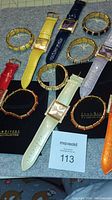 Photo showing 6 Joan Rivers Classics Collection watches with gold tone metallic faces and leather bands in red, ivory, yellow, lavender, navy, and orange. Accompanied by 6 coordinating gold tone enamel bangle bracelets with rhinestone accents.