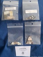 Four transparent ziplock bags each containing distinct jewelry pendant pieces labeled as 'Mother of Pearl Stainless Steel' and 'Abalone Onyx 925 Silver' on a blue surface next to a MaxSold auction card.