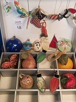 Display of various holiday ornaments including stained glass, hand painted wood, and glittered fruit shapes arranged in a box and hanging above.
