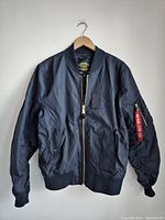 Full front view of the black Alpha Industries bomber jacket hanging on wooden hanger showing zipper closure and front pockets