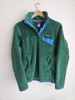 Front view of a green Patagonia Snap-T fleece pullover jacket with blue snap button placket and pocket flap, hanging on a wooden hanger.