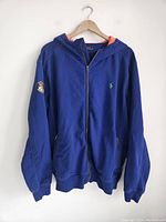 Blue Ralph Lauren Polo hoodie hanging on a hanger showing full frontal view, hood lined with orange fabric, full zipper, front pockets, ribbed cuffs and hem.