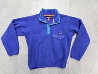 Full view of blue Patagonia Snap-T fleece pullover with orange trim and teal collar laid flat on concrete.