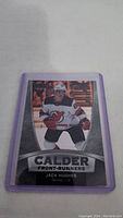 Photo of a Jack Hughes rookie hockey card from the Calder Front-Runners series, showing player in New Jersey Devils uniform encased in a protective purple plastic sleeve.