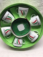 Top-down view of six white cappuccino cups and a saucer in a green box with 'cappuccino' text and red and green stripes on cups.