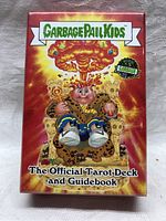 Front of sealed red box for Garbage Pail Kids official tarot deck and guidebook showing character sitting on throne with explosion background.