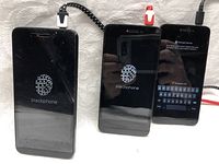 Three black Blackphone smartphones powering on with Blackphone logo on screens. One phone displays decrypt device password prompt. Phones connected to charging cables without chargers.