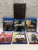Front view of six PS4 game cases with artwork visible, including Far Cry 4, Call of Duty Infinite Warfare, Assassin's Creed Origins, Wolfenstein The Old Blood, Doom, Bloodborne. Cases are arranged with a background featuring a textured dark item in the center.