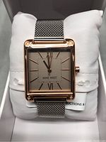 Close-up of the watch face showing the square rose gold-tone case, silver mesh band, and light dial with Roman numerals and stick markers, inside the Nine West box on a white cushion.