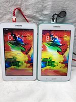 Pair of Samsung tablets powered on showing colorful lock screen with time and date, connected with charging cables (red and black).