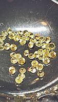 Close-up photo of many small round yellow sapphire stones with diamond cuts placed in a dark metal bowl, showing their bright yellow color and sparkle.