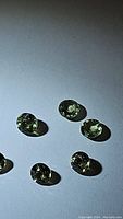 Seven natural light green round faceted sapphires about 3mm diameter each, laid out on white surface showing their color and clarity.