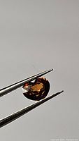 Close-up of heart cut burnt orange Cambodian zircon gemstone held between tweezers against neutral background showing clarity and facets.