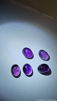Five oval cabochon cut amethyst stones showing deep purple color with visible inclusions, arranged on a white surface under direct light.