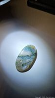 Photo of oval cabochon natural moss agate gemstone showing translucent body with internal green and blue moss-like markings under direct light.
