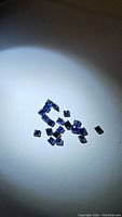 Small blue square cut sapphire gemstones spread on a white surface under light, showing blue color and square shapes.