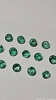 Photo showing thirteen small round calibrated natural emeralds, green in color with natural inclusions visible, arranged on a light surface.