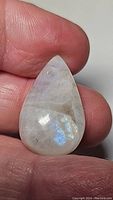 Front side of a pear-shaped rainbow moonstone cabochon with visible blue flash held between fingers for scale.