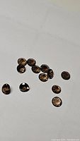 Twelve round faceted smokey quartz gemstones in deep brown color scattered on a white background, showing size and color consistency.