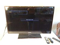 Front view of 40-inch Samsung TV turned on displaying 'Check signal cable' message with three remotes placed beside it.