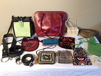 Full lot display of assorted bags, purses, wallets, basket container and belts on a white cloth surface against a beige wall.