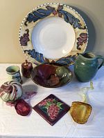 Full view of all items displayed together on white surface including ceramic platter, pitcher, glass lilies, sphere, lidded pot, decorative tile, and cup.