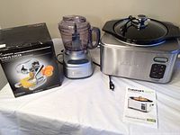 Overall view showing the Rabbit Citrus Juicer in boxed packaging, Cuisinart small food processor, and Cuisinart slow cooker with glass lid on a white tablecloth.