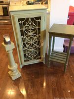 Photo showing the front of the green wooden cabinet with ornate metal door, small table, and wooden candlestick on a hardwood floor