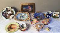 Full view of 7-piece porcelain collection including various bowls, creamers, and decorative items on a white surface with painting in background.
