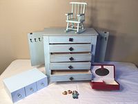 Front view of shabby chic wooden jewelry box with drawers open and side doors with hooks, small red leather jewelry box, tiny blue rocking chair on top, and painted earrings and pin laid out in front.