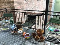 View of three outdoor chairs, various small ceramic and clay plant pots in front, watering can and garden gloves visible