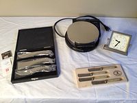 Full view of all four items in the lot: Laguiole cheese knife set, Phillipi cheese cutlery set, waffle griddle and small clock, displayed on white cloth.